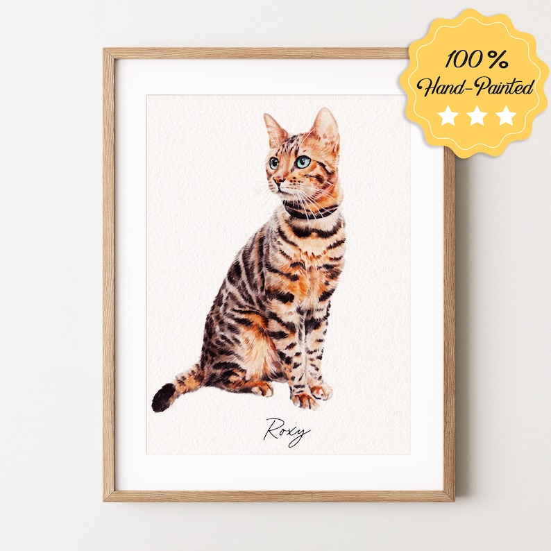 May include: A watercolor painting of a brown tabby cat with green eyes, sitting and looking to the left. The cat is named "Roxy" in cursive writing below the image. The painting is framed in a light wood frame.