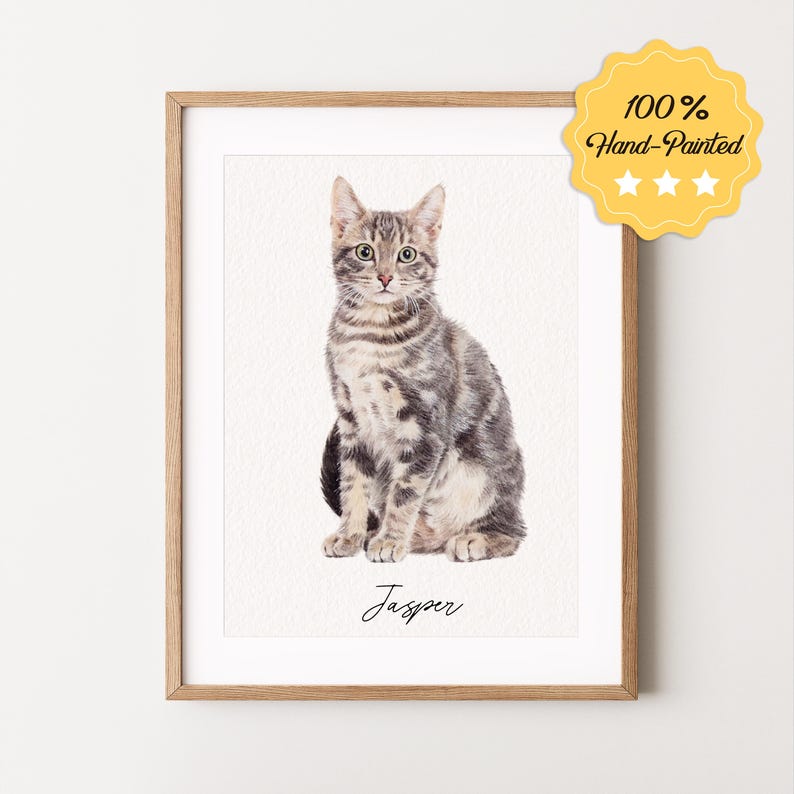 May include: A watercolor painting of a tabby cat named Jasper. The cat is sitting with its front paws tucked under its body and its tail curled around its legs. The painting is framed in a light wood frame.