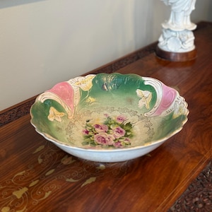 May include: An antique porcelain bowl with a scalloped edge. The bowl features a floral design with pink roses and green leaves in the center. The rim has pink and green accents with gold trim. A decorative piece for display.