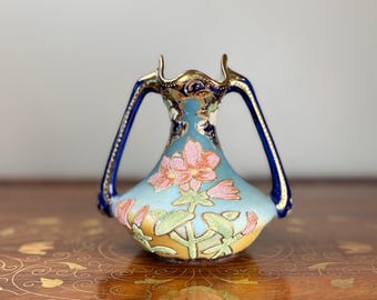 May include: A small ceramic vase with two handles, featuring a light blue and yellow gradient base decorated with raised pink flowers and green leaves. The neck is accented with dark blue and gold trim, and the rim has a scalloped edge design.