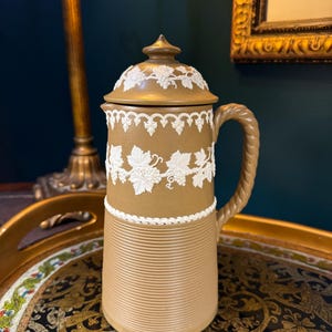 May include: A tan ceramic pitcher with a lid, featuring white grape vine detailing. The pitcher has a ribbed base and a braided handle. The pitcher is approximately 10 inches tall.