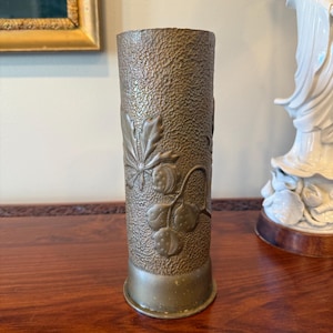 May include: A bronze-colored, cylindrical vase with a textured surface and a decorative relief of leaves and berries. The vase has a wide base and a slightly flared top. The design is likely inspired by nature and adds a touch of elegance to any space.