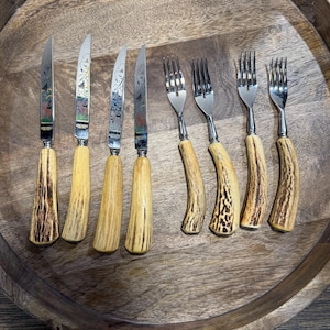 May include: A set of six knives and forks with decorative blades and tines. The knives have ornate designs on the blades and light brown handles. The forks have silver tines and light brown handles. The set is arranged on a wooden tray.