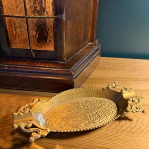 May include: A gold-colored, oval-shaped decorative tray with ornate floral and scrollwork patterns. The tray has a scalloped edge and decorative flourishes at each end. A wooden cabinet with a stained glass window is in the background.