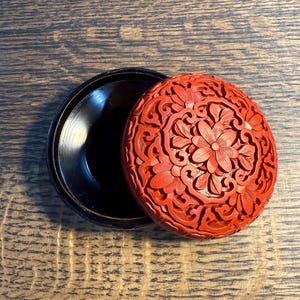 May include: A round, black container with a detached, red lid. The lid features a detailed floral carving design. The container is open, revealing its dark interior. The piece is set against a wooden surface.