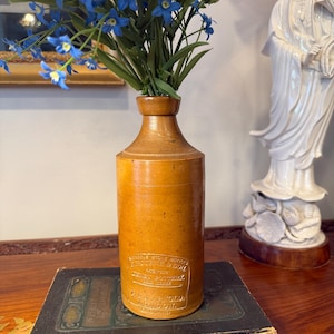 May include: A vintage, tan-colored stoneware bottle with a label, filled with blue flowers. The bottle is cylindrical with a narrow neck and sits on top of a stack of old books. The label reads "BOURNE & SON" and "LONDON".