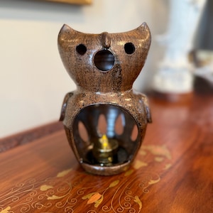 May include: A bronze-colored owl-shaped oil warmer. The owl has large eye holes and a circular opening for the mouth. The base has a large opening to hold a small candle. The warmer is sitting on a wooden surface.