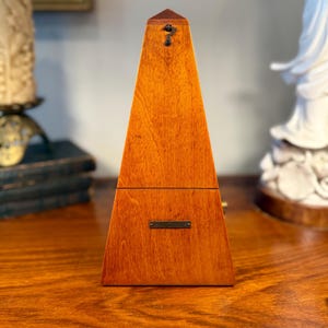 May include: A pyramid-shaped wooden metronome with a warm brown finish. The front features a small metal plate near the center and a metal hook near the top point. The device has a smooth, polished surface showing natural wood grain patterns.