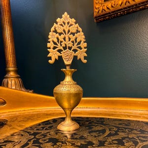 May include: A vintage brass perfume bottle with a decorative tree-shaped stopper. The bottle has an ornate design and sits on a tray with a black and gold floral pattern. The background is a dark teal color.