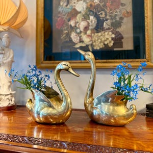 May include: Two polished brass swan-shaped planters, each featuring a hollow back designed to hold small arrangements. Both metallic figures are filled with vibrant blue flowers, showcasing a smooth, reflective surface and elegant, curved necks.