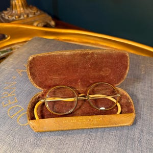 May include: Vintage round eyeglasses with a gold-tone frame and clear lenses, resting inside an open, brown leather case with a velvet lining. The case has a yellow strap and text that reads "R.O. Smith, M.D. Eye, Ear, Nose & Throat, Pulaski, Virginia."