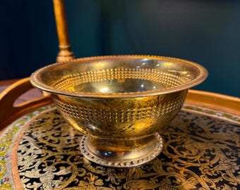 May include: A gold-colored metal bowl with a decorative, dotted pattern around the rim and body. The bowl sits on a small, silver-colored pedestal. The bowl is on a black and gold patterned tray.