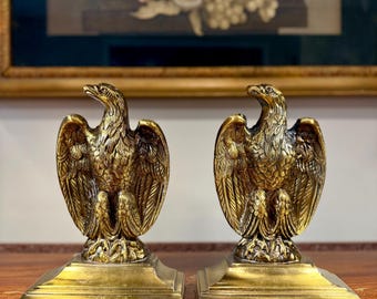 May include: Two antique brass eagle sculptures. Each eagle is perched on a square base with detailed feathering and a regal pose. The sculptures are displayed on a wooden surface with a floral painting in the background.