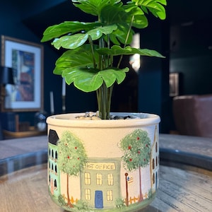 May include: A decorative ceramic planter with a hand-painted village scene, including a post office, trees, and houses. The planter holds a lush, green plant with large, textured leaves. The planter is approximately 6 inches in diameter.