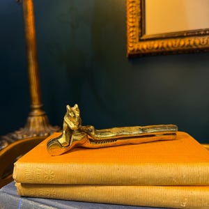May include: A vintage brass nutcracker with a fox head design. The nutcracker is resting on top of two orange books and a blue book. The background includes a lamp and a framed picture.