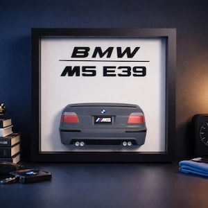 Könnte beinhalten: Gerahmtes Kunstwerk mit einer grauen BMW M5 E39 Heckansicht. Der schwarze Rahmen umgibt einen weißen Hintergrund mit dem BMW-Logo und dem Modellnamen. Ein kleines Modellauto, Bücher und Zubehör stehen auf dem Schreibtisch.