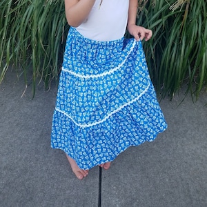 May include: A young girl wearing a white shirt and a blue and white tiered skirt with white lace trim. She is holding a white hydrangea flower in front of her face.