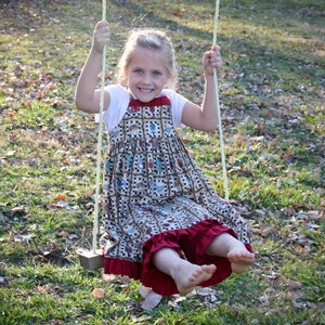 Może przedstawiać: Młoda dziewczyna o blond włosach uśmiecha się, siedząc na huśtawce. Ma na sobie brązową i białą sukienkę z czerwoną falbanką i białą koszulkę. Huśtawka jest wykonana z liny i drewna.
