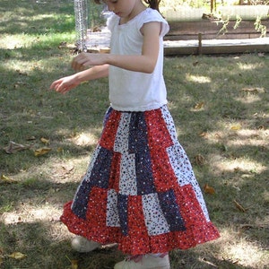 May include: A young girl wearing a white shirt and a red, white, and blue patchwork skirt. She is wearing white sneakers and has her hair pulled back in a ponytail with a red and white bow.