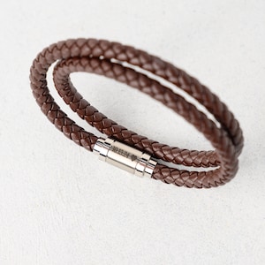May include: A brown braided leather bracelet with a silver clasp. The bracelet is made of two strands of braided leather. The clasp has the engraved text "10:13:24" with a heart symbol. The bracelet is approximately 8 inches in length.