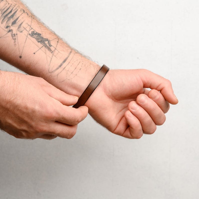 May include: A brown leather bracelet is being clasped on a wrist. The bracelet is a simple, thin band. The person's arm has a tattoo of a person and geometric shapes. The background is a plain white.