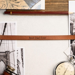 May include: A brown leather bracelet with gold-colored clasps, engraved with the text "Best Dad Ever." The bracelet is displayed on a white surface with black and white photographs and a stopwatch.
