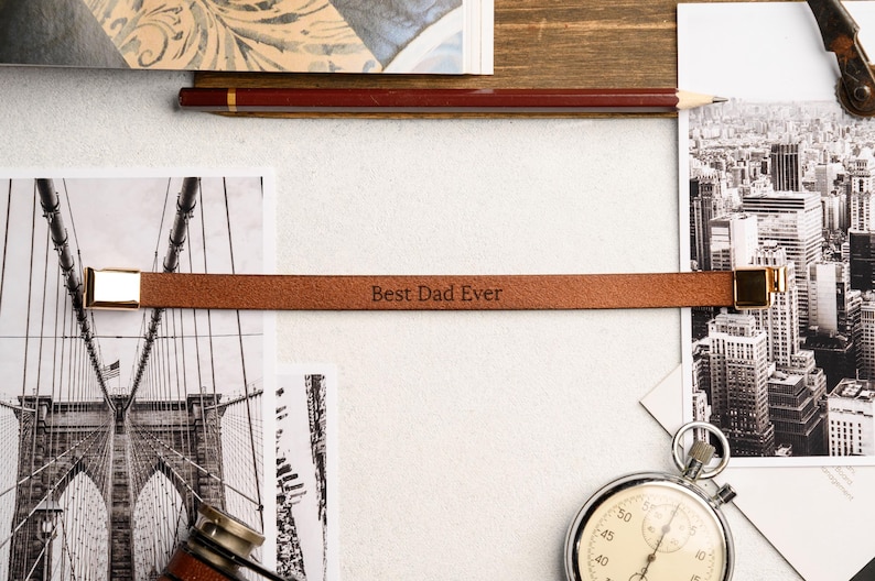 May include: A brown leather bracelet with gold-colored clasps, engraved with the text "Best Dad Ever." The bracelet is displayed on a white surface with black and white photographs and a stopwatch.