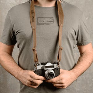 May include: A person wearing a gray t-shirt with the text "John Miller" and "freedom mode" printed on it. The person is wearing a brown leather camera strap and holding a vintage camera.