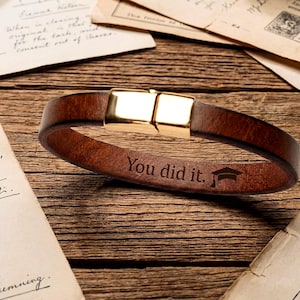 Pode incluir: Uma pulseira de couro castanho com fecho dourado. A pulseira é gravada com as palavras "You did it." e um símbolo de chapéu de formatura. A pulseira é exibida em uma superfície de madeira com papéis vintage.