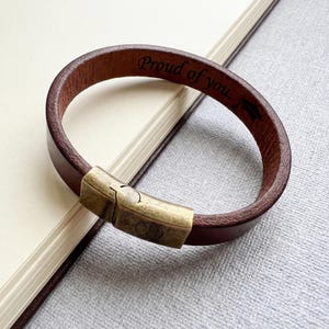 May include: Brown leather bracelet with a bronze-colored clasp. The bracelet is engraved with the words "Proud of you." The bracelet is resting on a notebook.