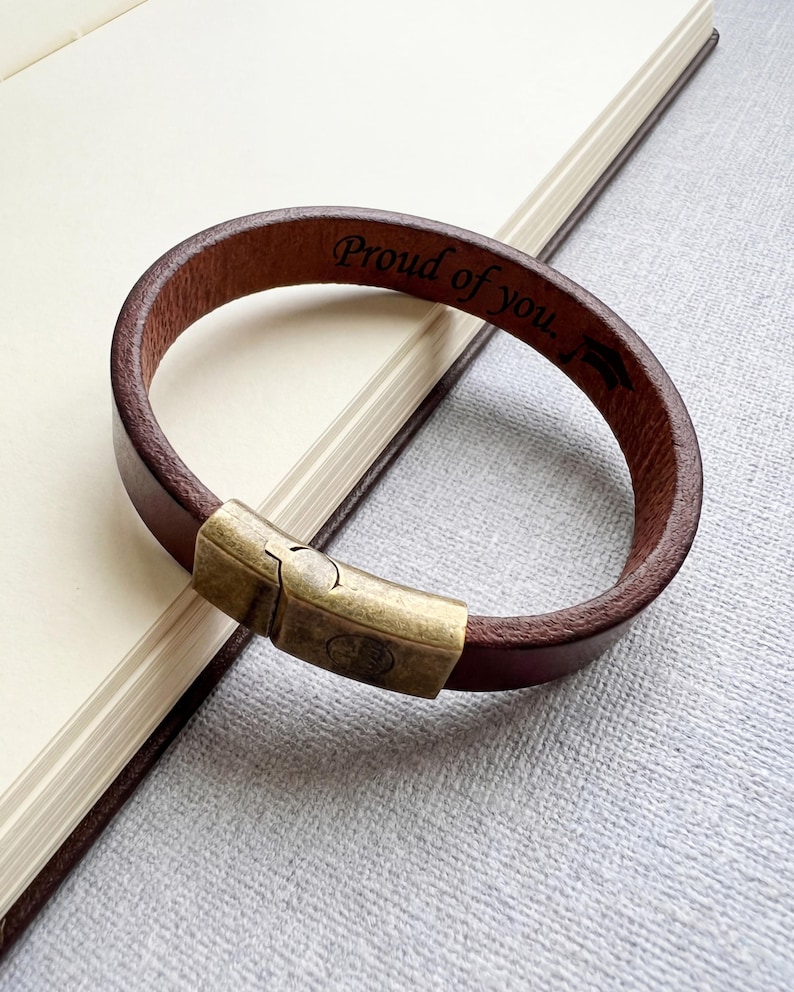 May include: Brown leather bracelet with a bronze-colored clasp. The bracelet is engraved with the words "Proud of you." The bracelet is resting on a notebook.
