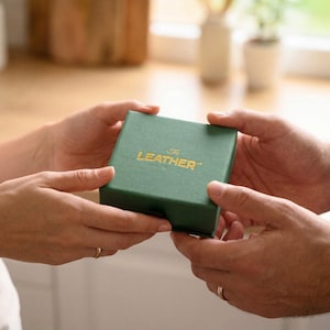 May include: A green box with the text "The Leather" in gold lettering is held between two sets of hands. The box is square and appears to be made of a textured material. The background is blurred, suggesting a domestic setting.