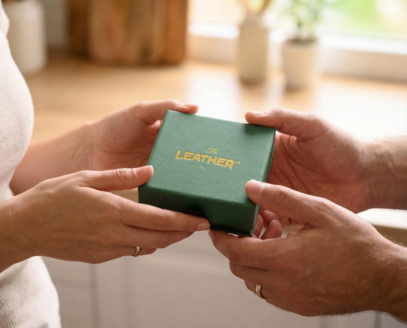 May include: A green box with the text "The Leather" in gold lettering is held between two sets of hands. The box is square and appears to be made of a textured material. The background is blurred, suggesting a domestic setting.