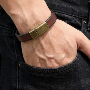 May include: A brown leather bracelet with a rectangular gold-colored clasp. The bracelet is worn on a wrist, with a dark denim background.