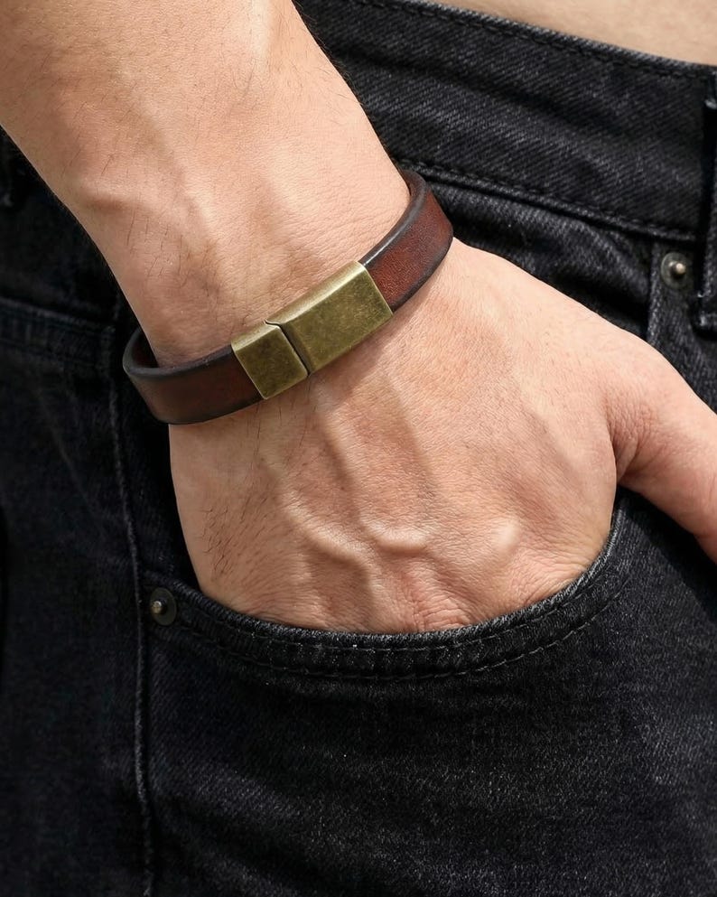 May include: A brown leather bracelet with a rectangular gold-colored clasp. The bracelet is worn on a wrist, with a dark denim background.