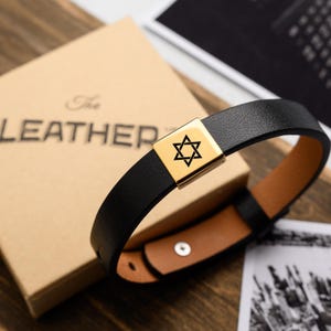 May include: Black leather bracelet with a gold-coloured clasp featuring a Star of David symbol. The bracelet is displayed next to a tan box with the word "LEATHER" printed on it. The bracelet has a brown interior.