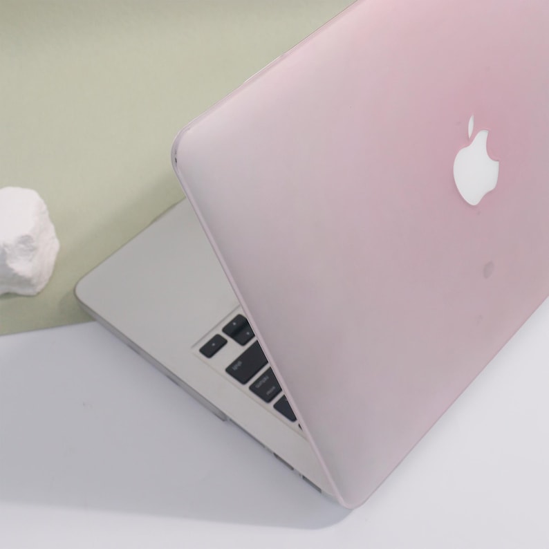 May include: A pink laptop case on a silver laptop. The Apple logo is visible on the lid. The keyboard is black with white lettering. The background is a light green and white.