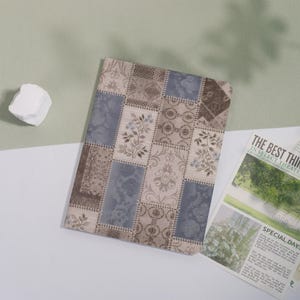May include: A rectangular notebook with a patchwork design in shades of beige, brown, and blue. The cover features floral and damask patterns. A white rock and a newspaper with the headline "THE BEST THING" are also in the image.