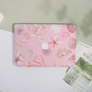May include: Pink laptop case with a watercolour design of seashells, seahorses, coral, and flowers. The case has a light pink background and a white Apple logo. The design includes various shades of pink and coral.