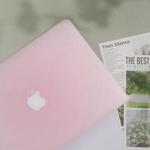 May include: A light pink laptop with the Apple logo on the lid. The laptop is on a white surface with printed materials, including the text "Then Silence" and "THE BEST IN SELECT". The background is a light green fabric.