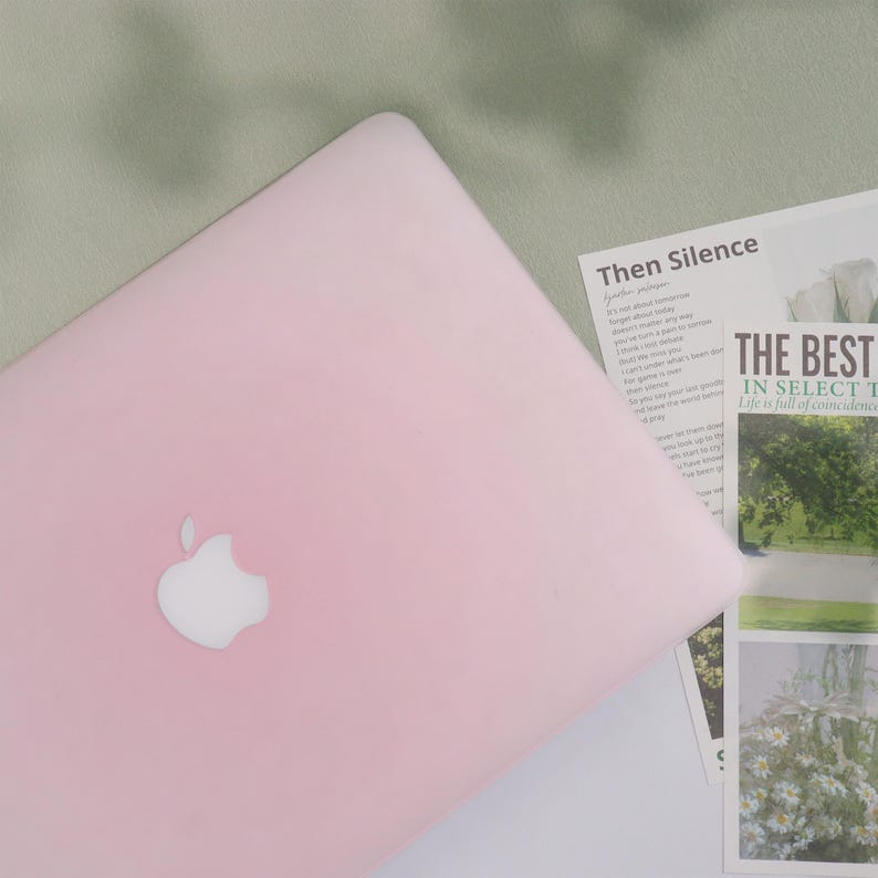 May include: A light pink laptop with the Apple logo on the lid. The laptop is on a white surface with printed materials, including the text "Then Silence" and "THE BEST IN SELECT". The background is a light green fabric.