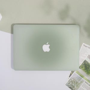 May include: A pale green Apple laptop with the Apple logo in the centre. The laptop is on a white and light green surface. The laptop is closed. There are some printed papers to the right of the laptop.