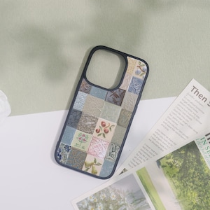 May include: A phone case with a patchwork design of various square tiles in shades of blue, beige, and gray. Some tiles feature floral and fruit motifs. The case has a black border and is lying on a white surface.