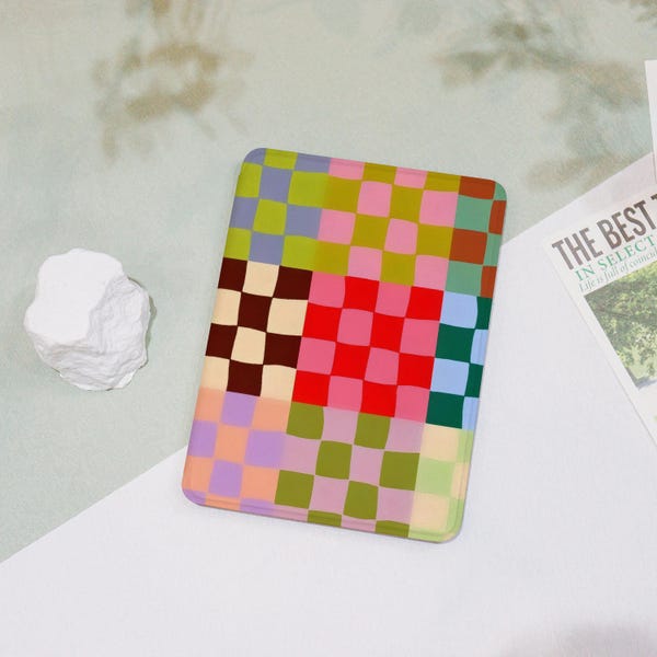 Puede incluir: Una funda rectangular con un colorido patrón de tablero de ajedrez en tonos verdes, rosas, rojos, marrones y azules. La funda está sobre una superficie blanca y verde claro. A la izquierda hay un objeto blanco en forma de roca.