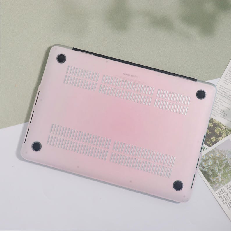 May include: A clear, pink MacBook Pro case. The case has a series of ventilation slots and four black rubber feet. The text "MacBook Pro" is visible on the case. The case is designed to protect the laptop from scratches and other damage.