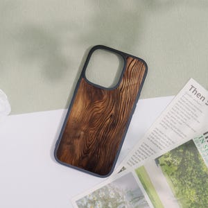 May include: A phone case with a dark wood grain design. The case has a dark blue border and a natural wood pattern. The phone case is lying on a white surface with a magazine and a green background.
