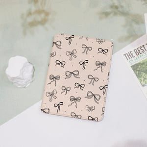 May include: A light pink tablet case featuring a repeating pattern of black bows. The bows vary in size and style, creating a playful and feminine design. The case is rectangular and appears to be made of a smooth material.