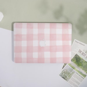 May include: A pink and white gingham pattern laptop case with the Apple logo in the center. The case is on a white and green background. The words "Then Silence", "The Best In Select Thea", and "Special Days" are visible on the papers.
