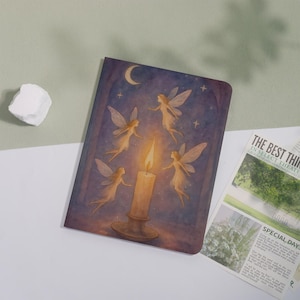 Könnte beinhalten: Ein Notizbuch mit einer Fantasy-Illustration. Das Cover zeigt eine Kerze mit Flamme, umgeben von Feen, einem Mond und Sternen. Die Farbpalette umfasst Lila-, Gold- und Beigetöne. Das Notizbuch liegt auf einer weißen Oberfläche.