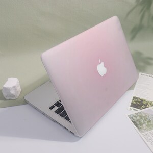 May include: A pink laptop with a white Apple logo on the lid is open on a white surface. The keyboard is visible, and a small white rock and some papers are nearby. The laptop has a silver base.
